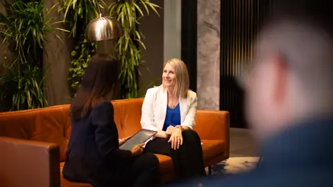 Two female employees having a conversation sitting on orange sofa, smiling in a plant-filled office.