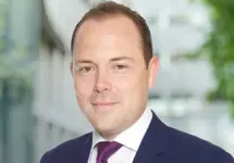Portrait of Adam Bywater, Director, standing outside in front of an outdoor urban setting, featuring modern architectural elements with clean lines. There are hints of greenery, suggesting some trees or plants.