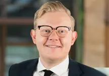 Ben Carter, Principal Consultant, UK, outside in front of a blurred brown background featuring a building and mix of natural and urban elements.