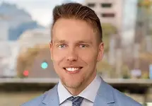 Portrait of Nicholas Rita, Associate Director, standing outside featuring an urban cityscape with tall buildings and a mix of architectural styles. The scene includes some trees with autumn foliage, adding a touch of color. 