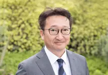Portrait of Toshio Kosuge, Director, Clean Energy and Transmission, Japan, standing outside against green bushes, in grey suit smiling.